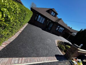 Tilted view of a brick house with a steep tiled roof, next to a tall green hedge along a paved driveway under a clear blue sky.